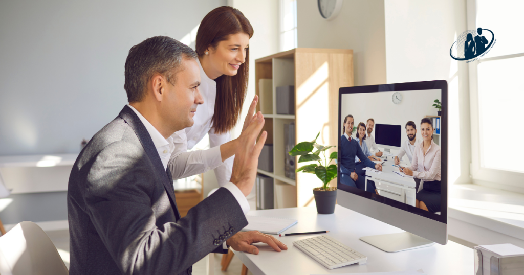 Two professionals wave at their offshore virtual team during a video call on a desktop monitor. The virtual team is seen smiling and seated in a well-lit office.